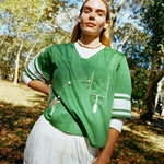 Woman wearing a green sports jersey and white shorts standing outdoors with trees in the background.