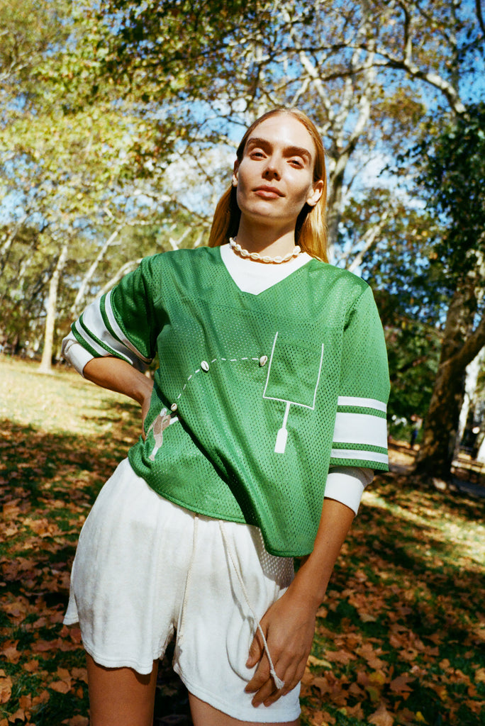 Woman wearing a green sports jersey and white shorts standing outdoors with trees in the background.