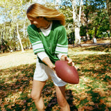 A woman wearing a green mesh football-style top with cowrie shells forming a football graphic, white shorts, and white sneakers while holding a football.