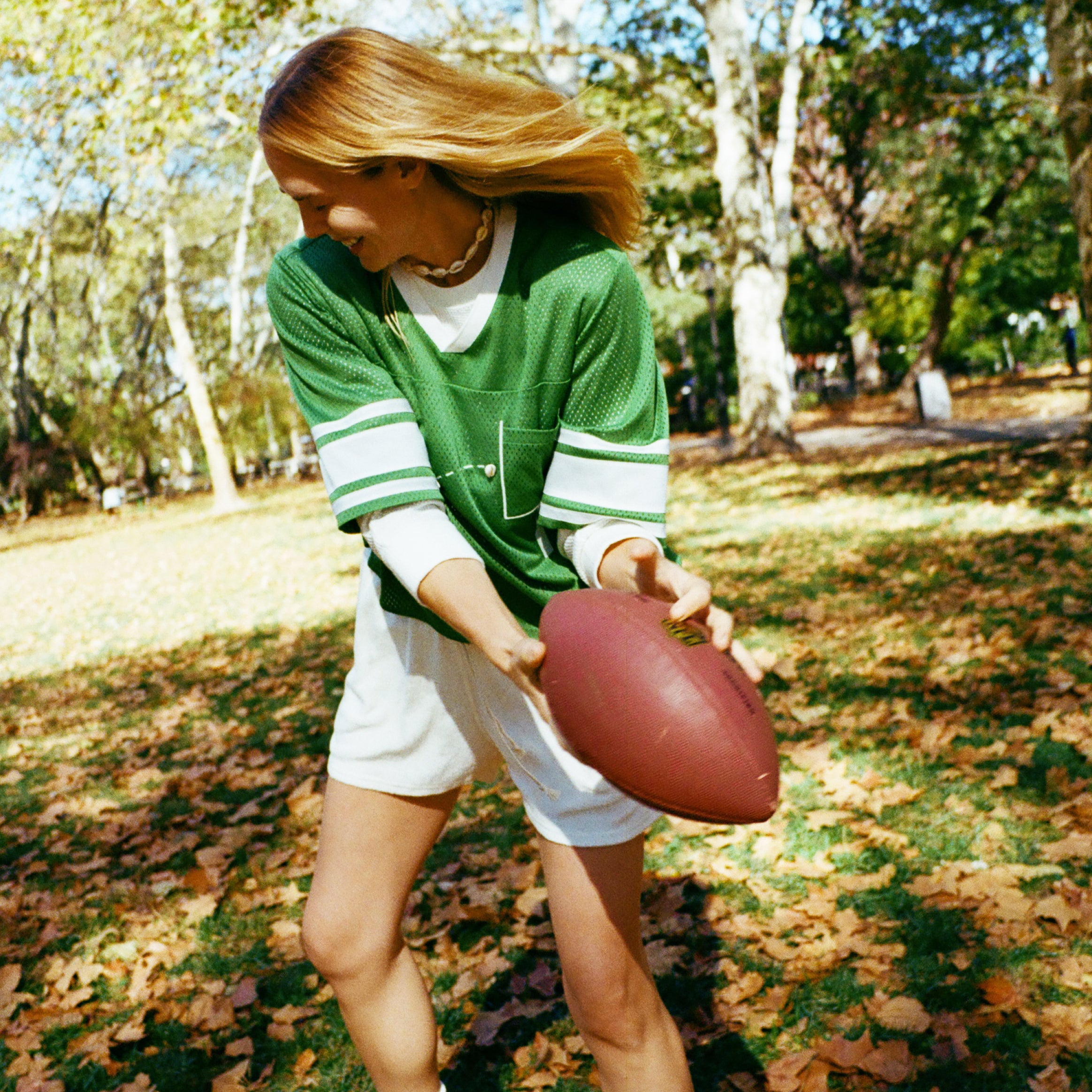A woman wearing a green mesh football-style top with cowrie shells forming a football graphic, white shorts, and white sneakers while holding a football.