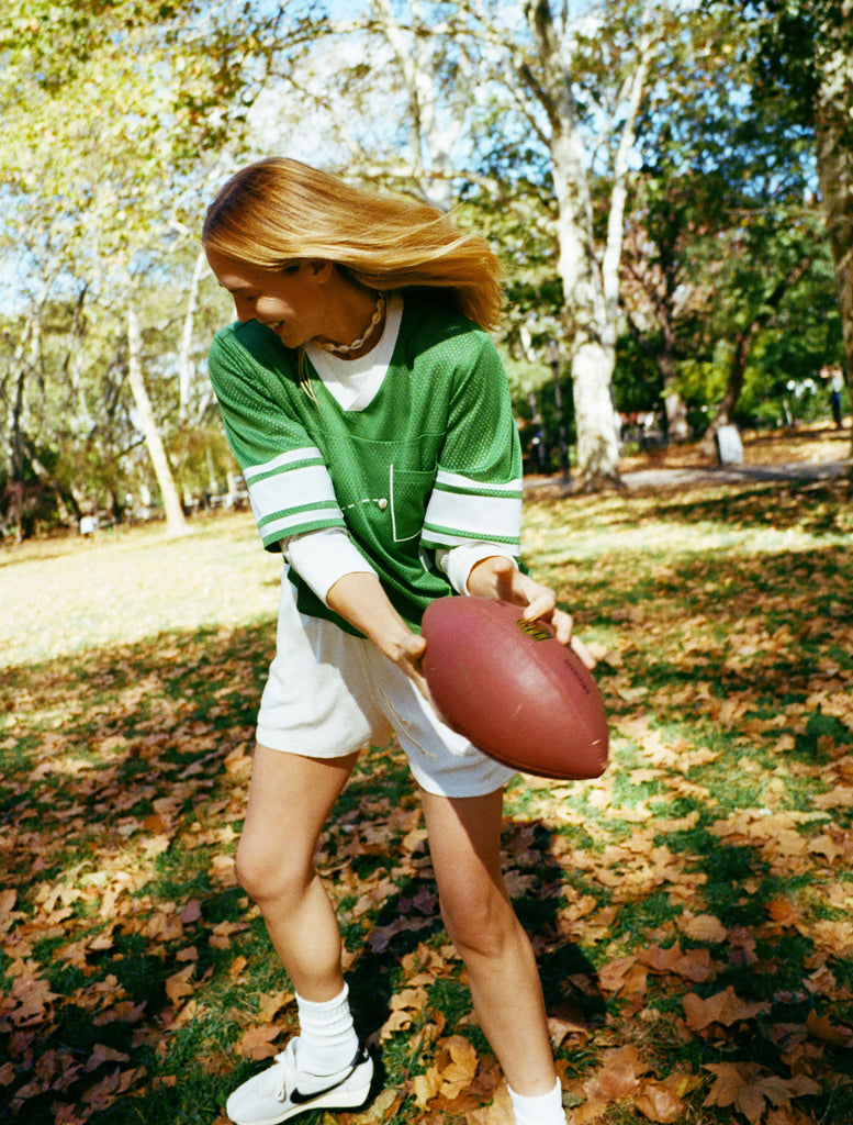 A woman wearing a green mesh football-style top with cowrie shells forming a football graphic, white shorts, and white sneakers while holding a football.