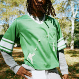A man wearing a green mesh football-style crop top with cowrie shells forming a football graphic, layered over a gray long-sleeve shirt and paired with gray athletic shorts.