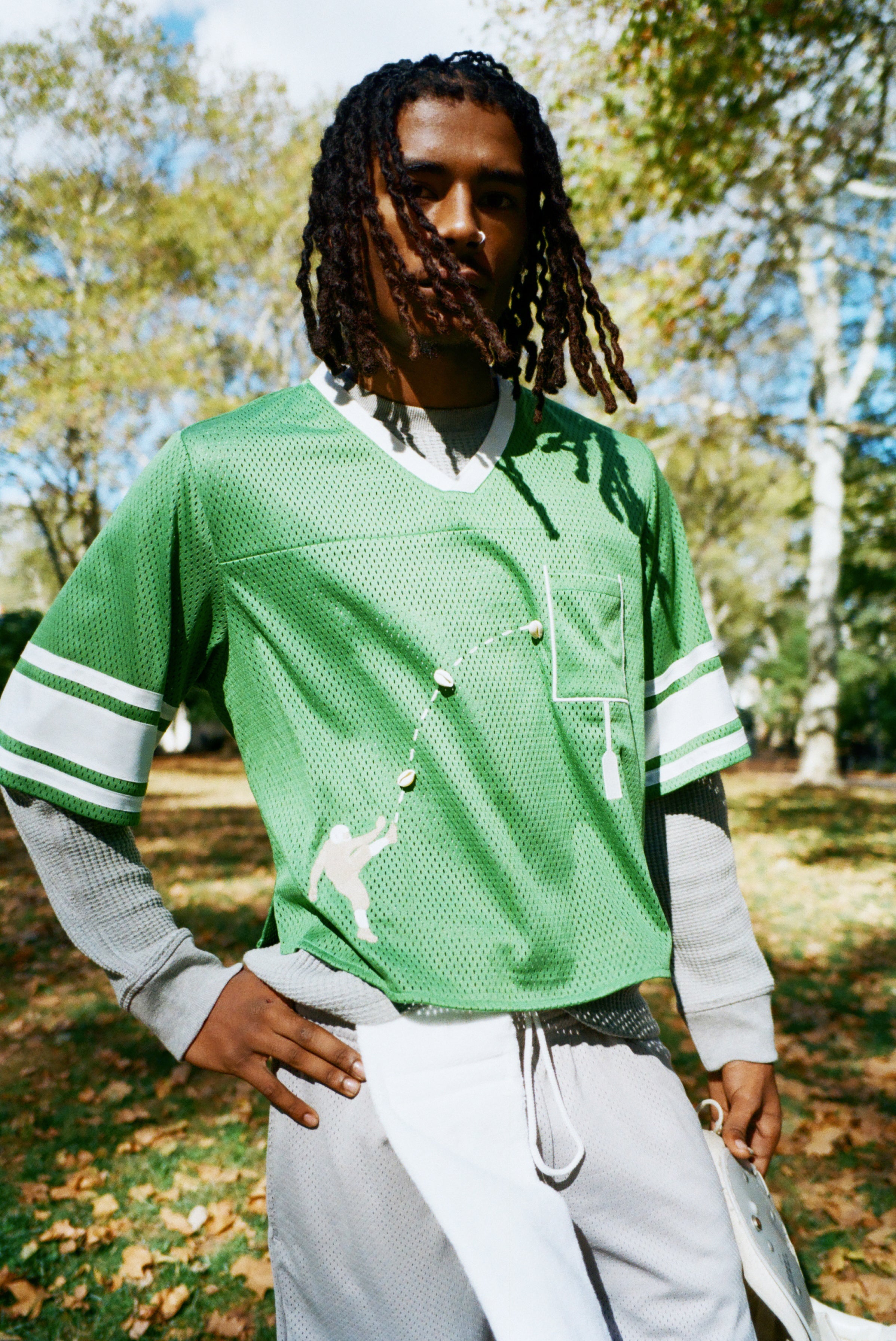 A man wearing a green mesh football-style crop top with cowrie shells forming a football graphic, layered over a gray long-sleeve shirt and paired with gray athletic shorts.