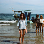 Three people stand in shallow ocean water with a small fishing boat anchored behind them. In the foreground, a woman faces the camera wearing a light blue button-up shirt featuring embroidered sand dollars and shell details, styled like a playful bikini top. In the background, a man in a black shirt and matching shorts stands beside a woman in a cream-colored shirt and sun hat.