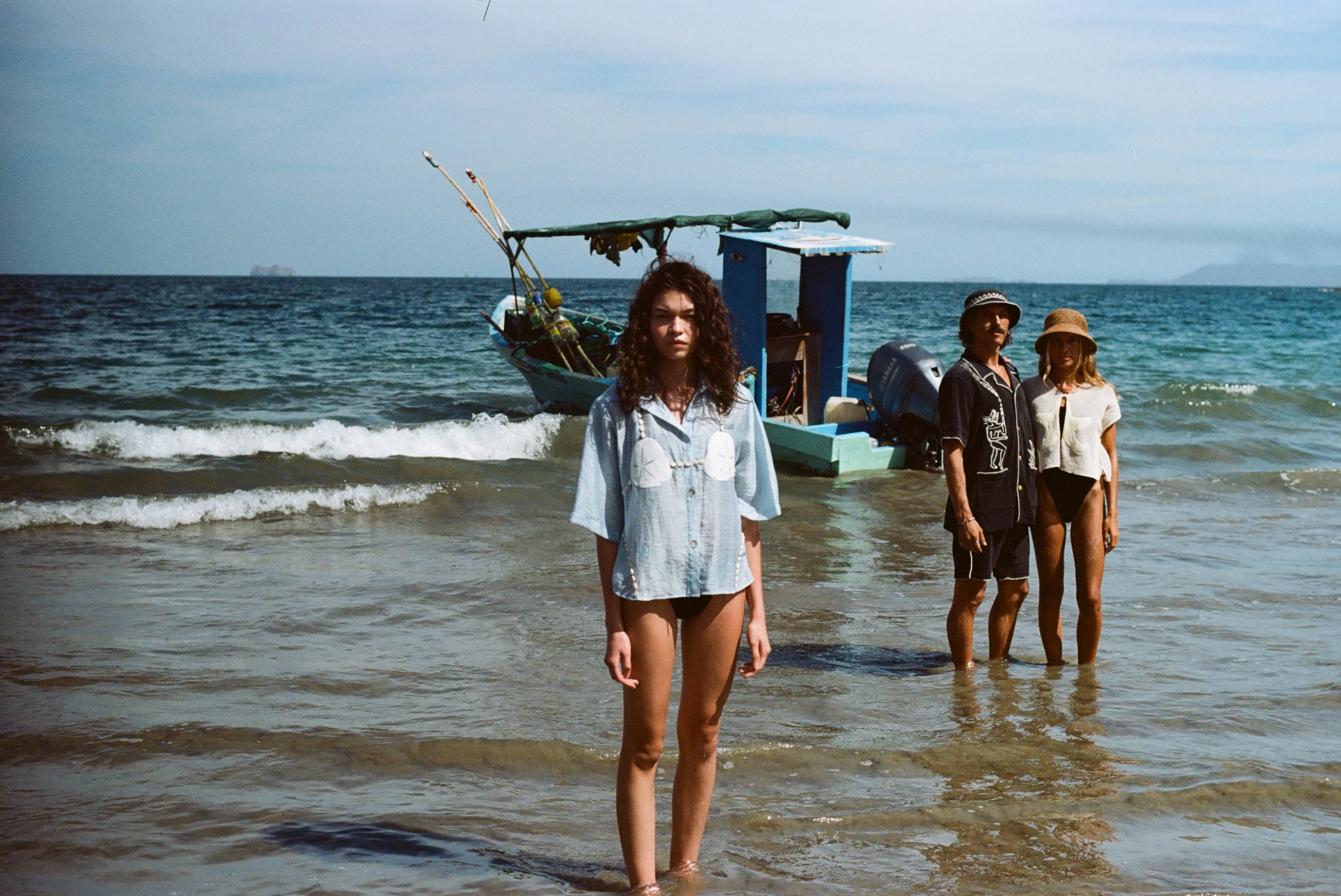 Three people stand in shallow ocean water with a small fishing boat anchored behind them. In the foreground, a woman faces the camera wearing a light blue button-up shirt featuring embroidered sand dollars and shell details, styled like a playful bikini top. In the background, a man in a black shirt and matching shorts stands beside a woman in a cream-colored shirt and sun hat.