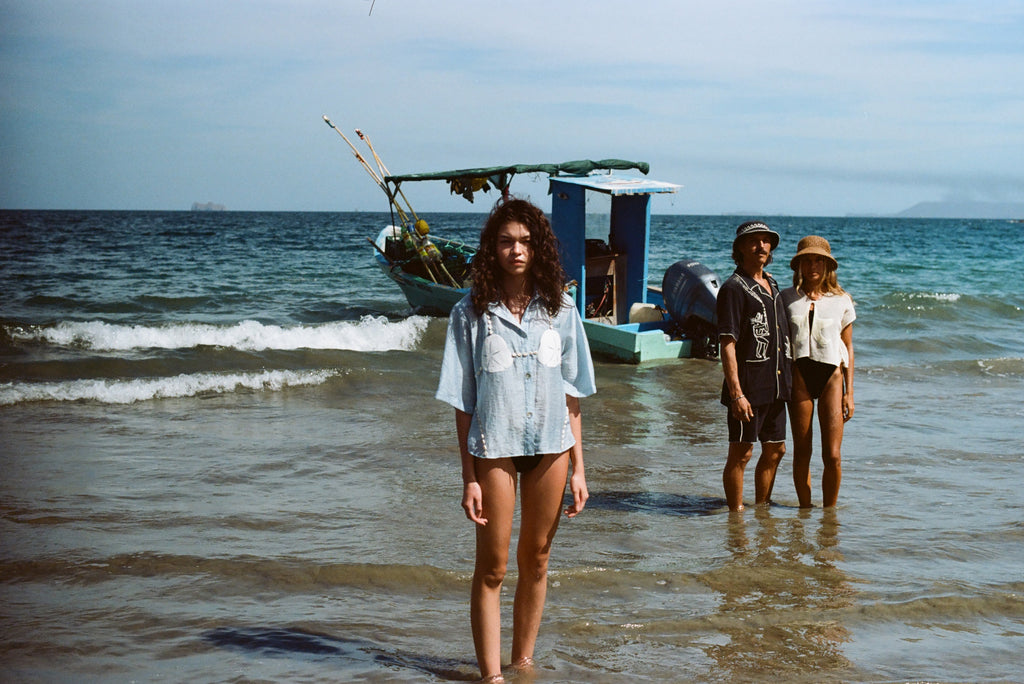 Three people stand in shallow ocean water with a small fishing boat anchored behind them. In the foreground, a woman faces the camera wearing a light blue button-up shirt featuring embroidered sand dollars and shell details, styled like a playful bikini top. In the background, a man in a black shirt and matching shorts stands beside a woman in a cream-colored shirt and sun hat.
