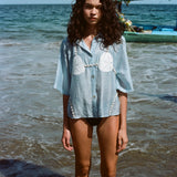 Image of woman in same light blue and dollar bikini shirt, standing ankle deep in the ocean, with a swim suit underneath.