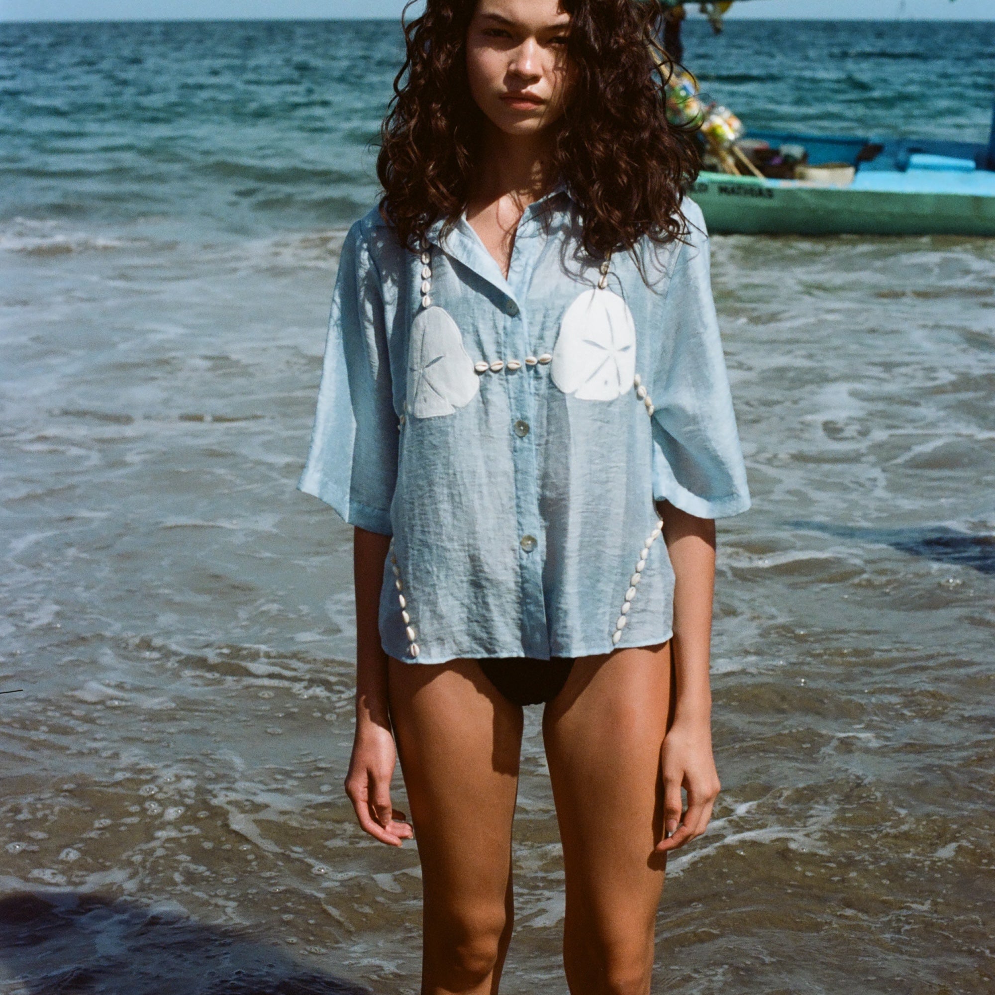 Image of woman in same light blue and dollar bikini shirt, standing ankle deep in the ocean, with a swim suit underneath.