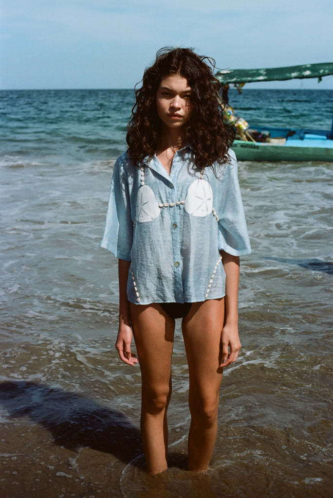 Image of woman in same light blue and dollar bikini shirt, standing ankle deep in the ocean, with a swim suit underneath.