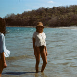 Woman standing ankle deep in ocean, wearing 'Starfish Strut' Hand Towel Top with a black swimsuit underneath and a straw hat.