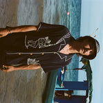 Sideways image of male model with longer hair and a mustache wearing the full 'Agua Magica' Cabana Set while standing ankle deep in the ocean. 