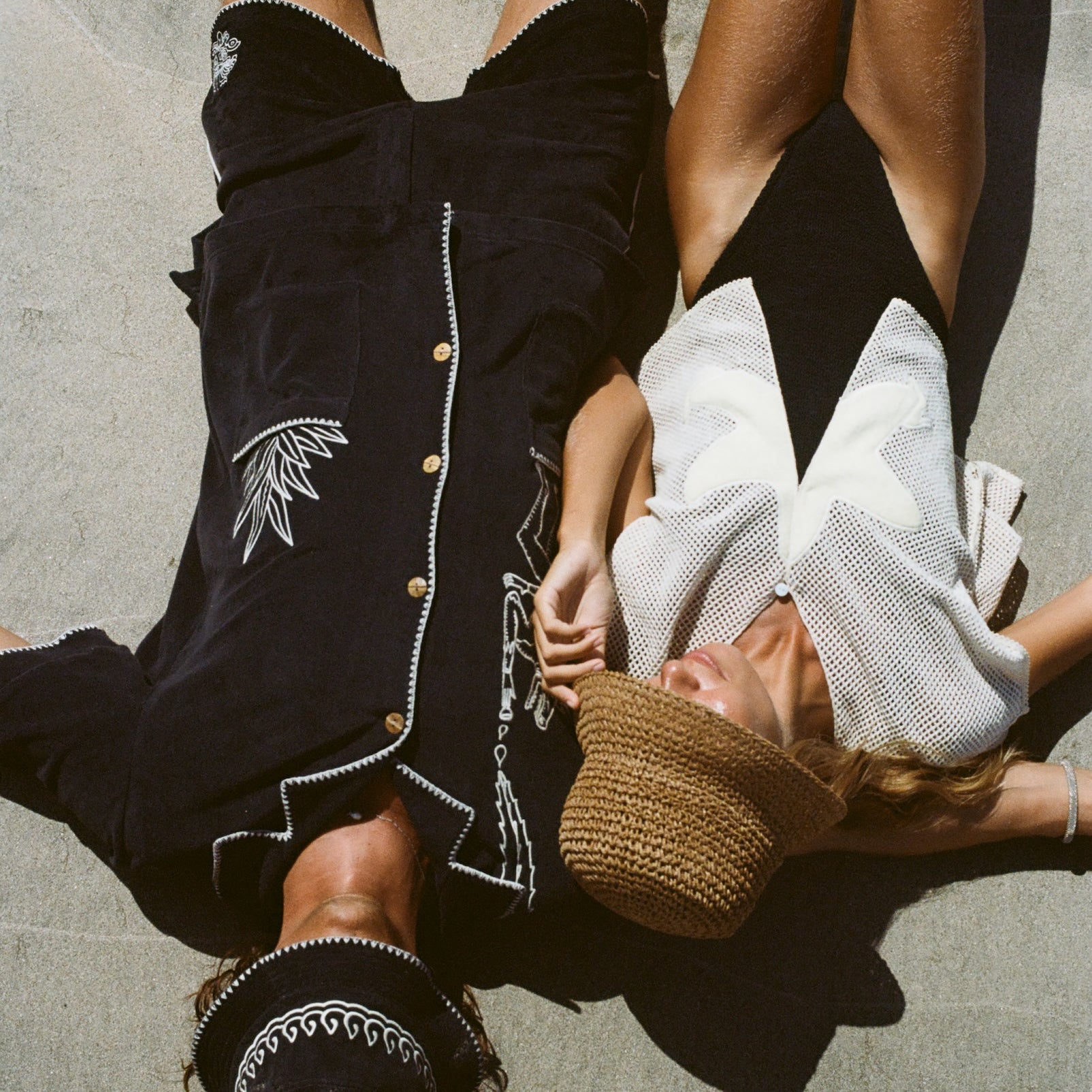 Two people lying on a beach. The man on the left wears a black outfit with white embroidered detailing and a matching hat, while the woman on the right wears the 'Starfish Strut' Hand Towel Top with a black swimsuit and a straw hat covering her face.