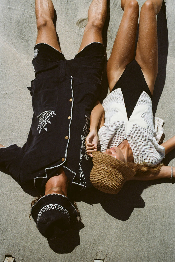 Two people lying on a beach. The man on the left wears a black outfit with white embroidered detailing and a matching hat, while the woman on the right wears the 'Starfish Strut' Hand Towel Top with a black swimsuit and a straw hat covering her face.