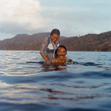 A woman and man smile while partially submerged in ocean water. The woman wears the blue shirt with oversized sand dollar and shell embroidery, playfully resting her hands on the man's shoulders. The man, in a floral print shirt, looks up with a big grin.
