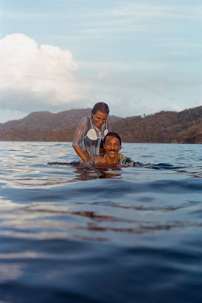 A woman and man smile while partially submerged in ocean water. The woman wears the blue shirt with oversized sand dollar and shell embroidery, playfully resting her hands on the man's shoulders. The man, in a floral print shirt, looks up with a big grin.