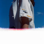 Woman wearing a white short-sleeve shirt with an embroidered seagull holding a fry on the sleeve, standing against a clear blue sky with light film burn at the bottom of the image.