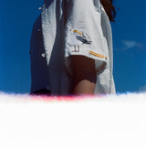 Woman wearing a white short-sleeve shirt with an embroidered seagull holding a fry on the sleeve, standing against a clear blue sky with light film burn at the bottom of the image.
