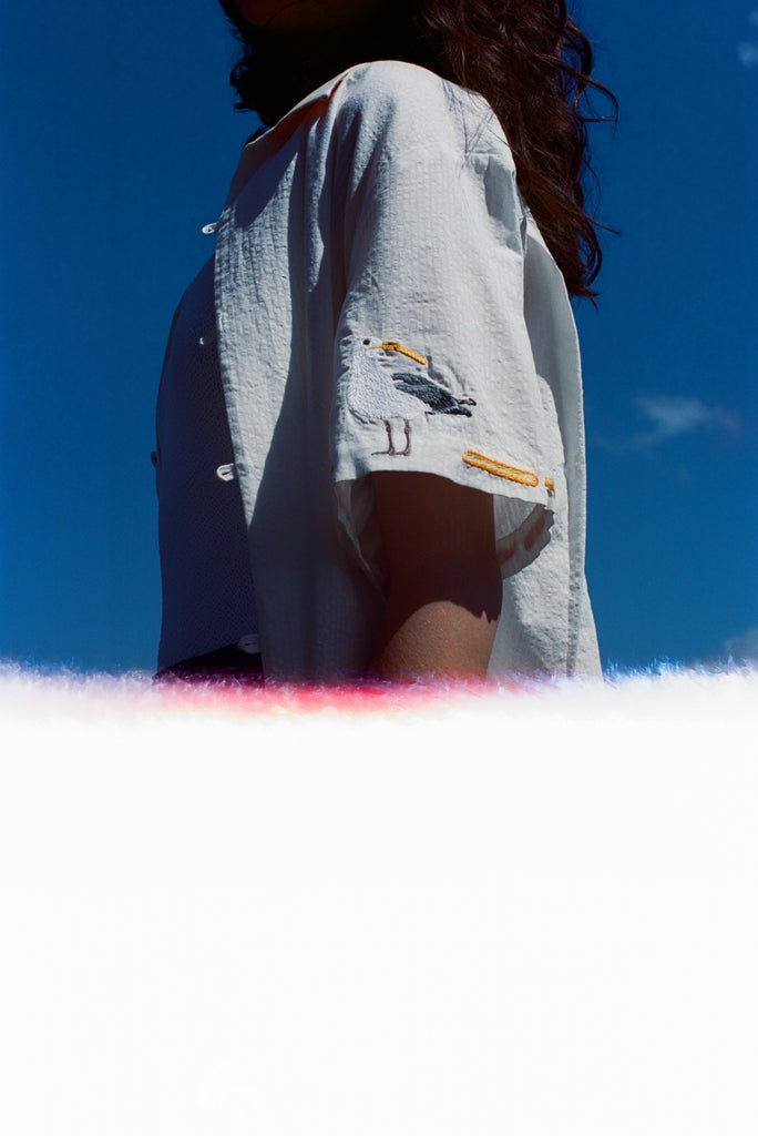 Woman wearing a white short-sleeve shirt with an embroidered seagull holding a fry on the sleeve, standing against a clear blue sky with light film burn at the bottom of the image.