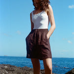 A female model stands on a rock at the beach, wearing the brown linen shorts and a white tank top. She has her hands in the pockets. 