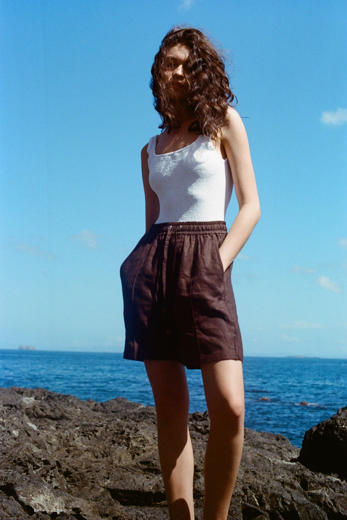 A female model stands on a rock at the beach, wearing the brown linen shorts and a white tank top. She has her hands in the pockets. 