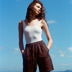 Female mode posing at the beach wearing a white tank top and the brown linen shorts, staring off to the left side as the sun hits her face.