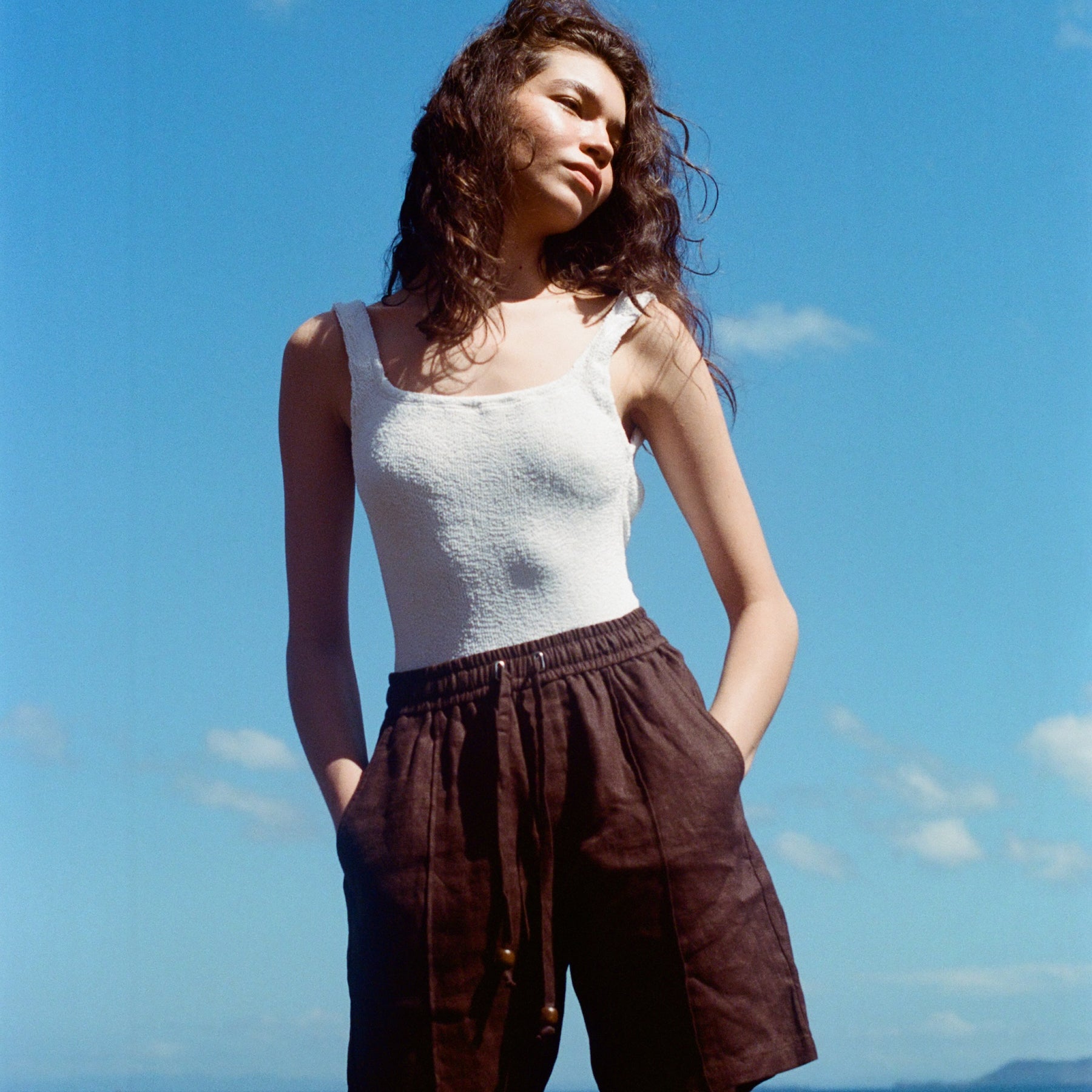 Female mode posing at the beach wearing a white tank top and the brown linen shorts, staring off to the left side as the sun hits her face.