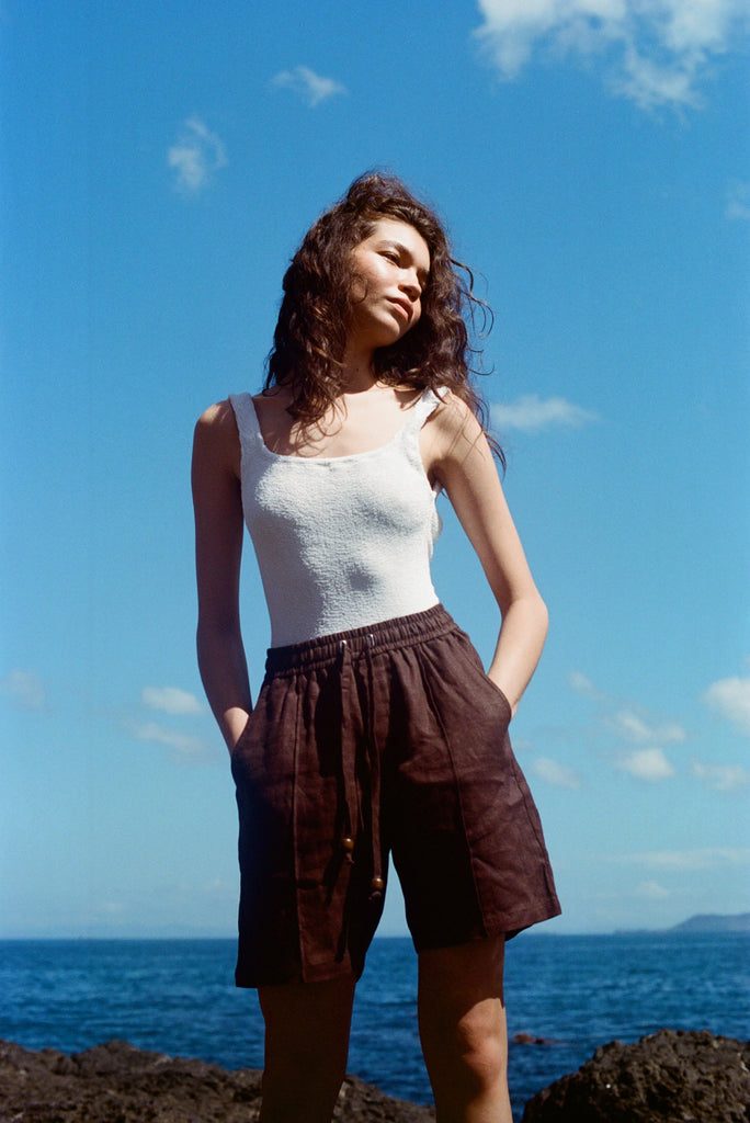 Female mode posing at the beach wearing a white tank top and the brown linen shorts, staring off to the left side as the sun hits her face.