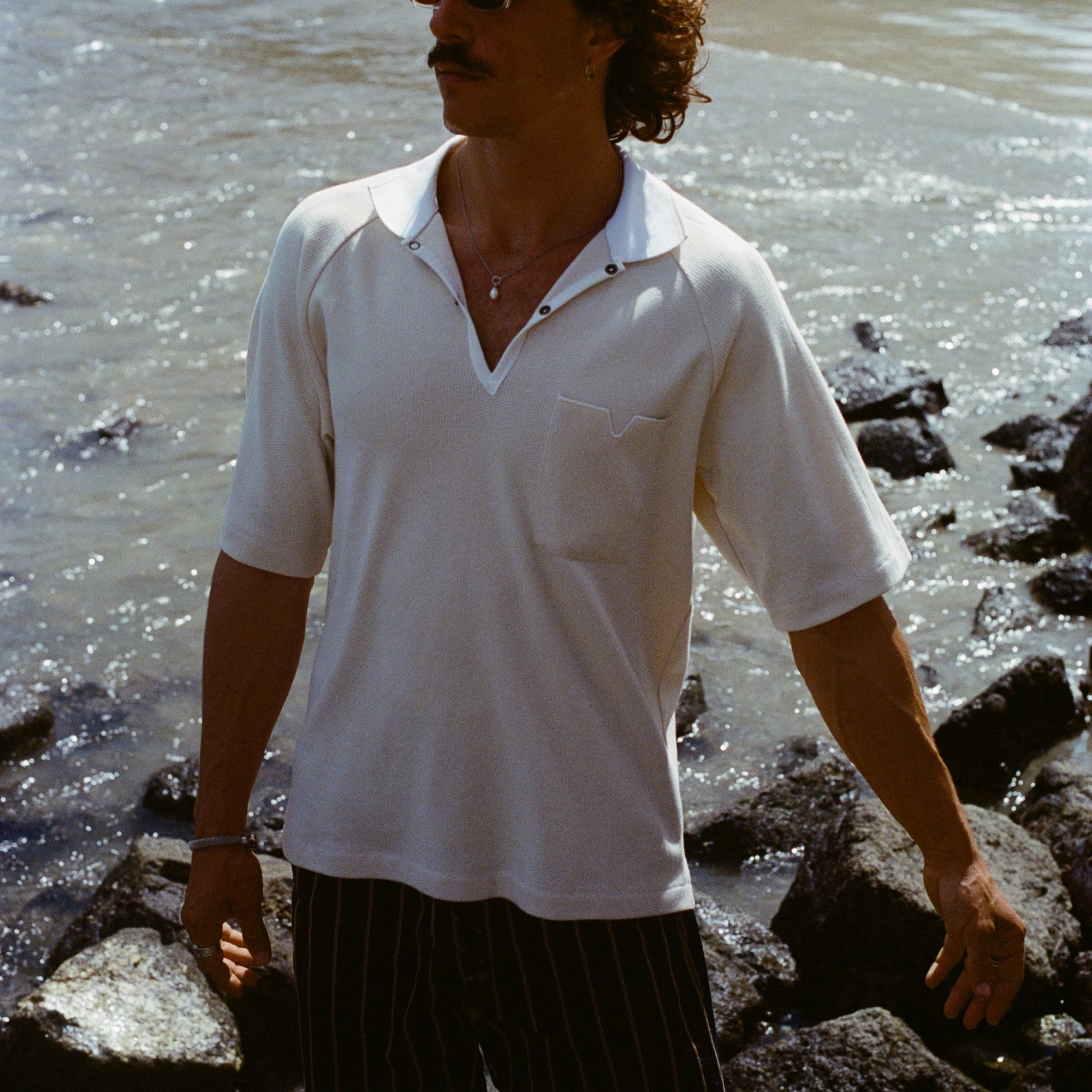 Man standing on rocky beach, wearing the beige mesh 'TomPolo' and striped shorts and looking off camera.