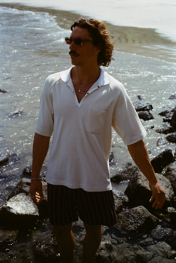 Man standing on rocky beach, wearing the beige mesh 'TomPolo' and striped shorts and looking off camera.