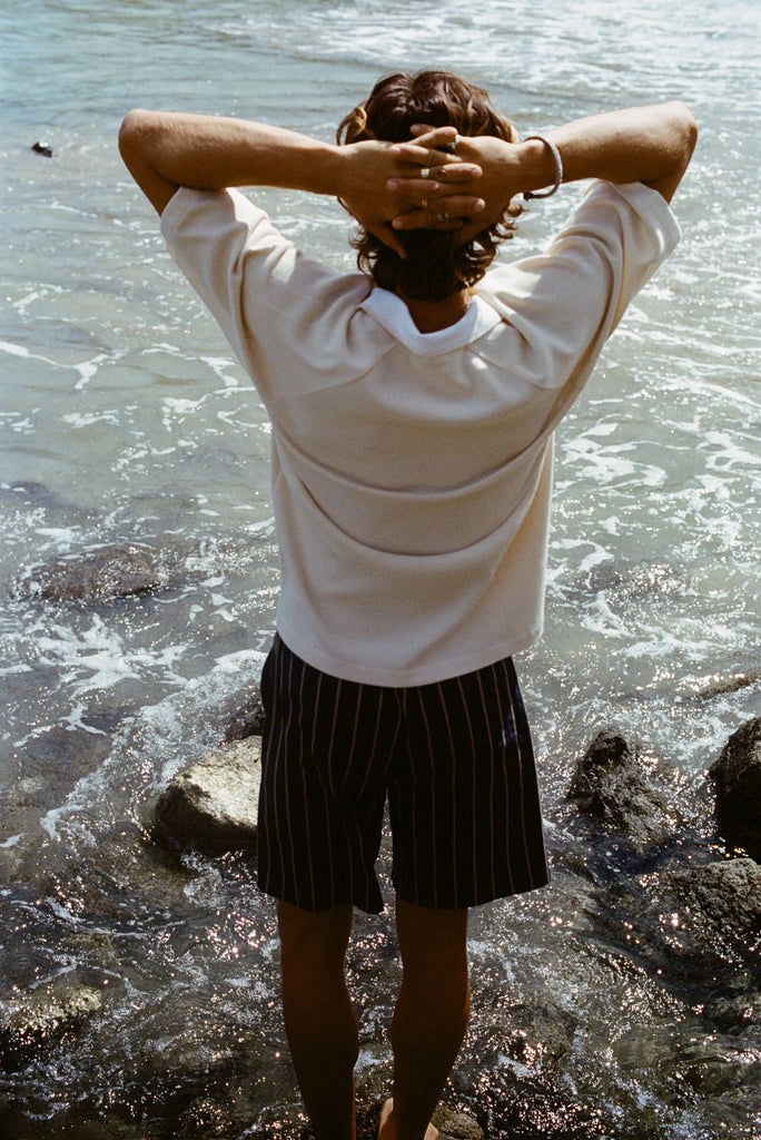 Man standing on shoreline with his back to us and his arms behind his head. He is wearing our beige mesh 'TomPolo' and our Easy Going Shorts in Navy Jacquard stripe. 