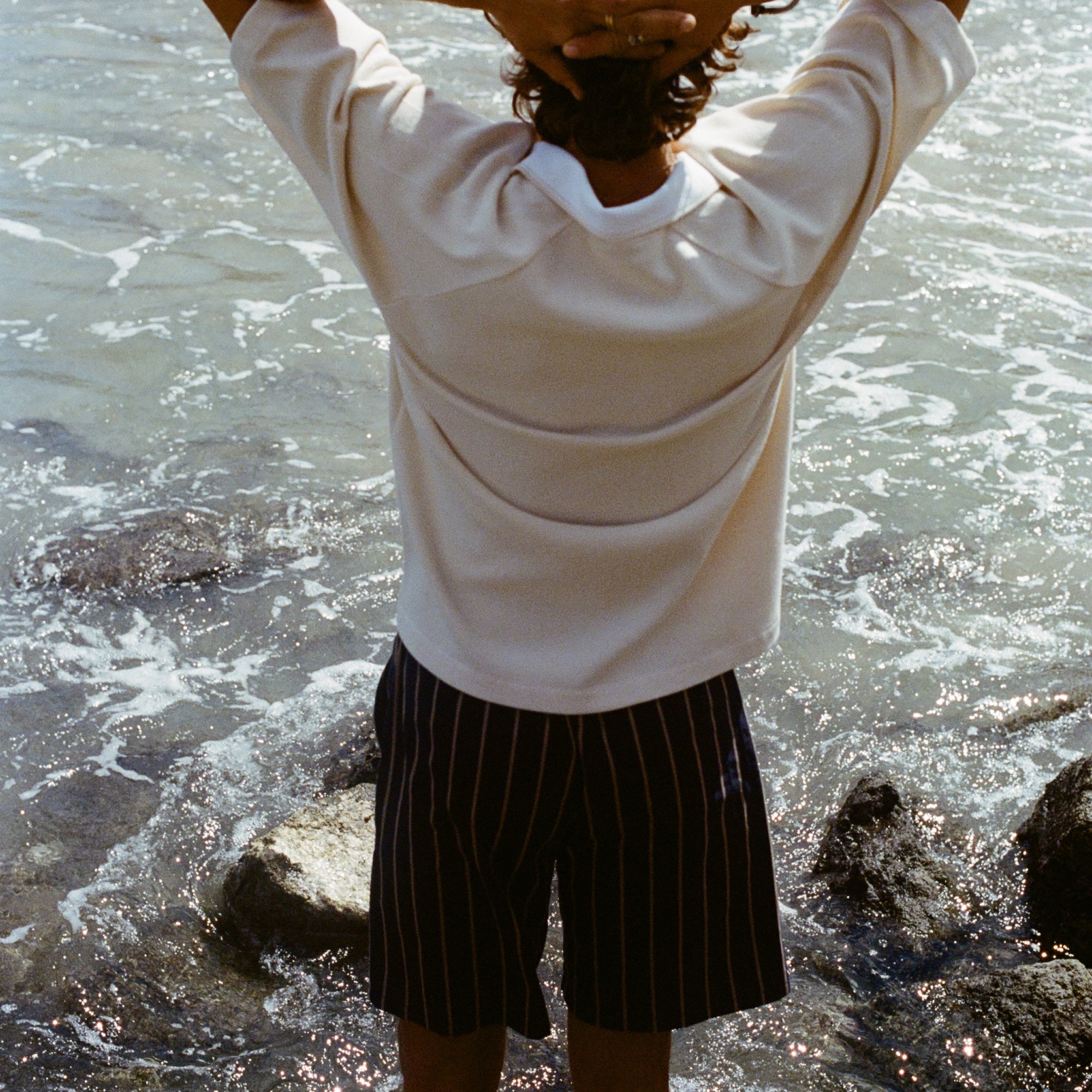 Image of a man's backside, wearing the beige mesh 'TomPolo' with striped shorts and has his hands behind his head. 
