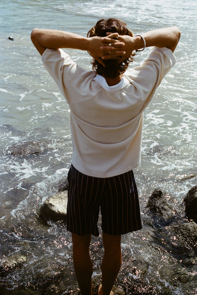 Image of a man's backside, wearing the beige mesh 'TomPolo' with striped shorts and has his hands behind his head. 