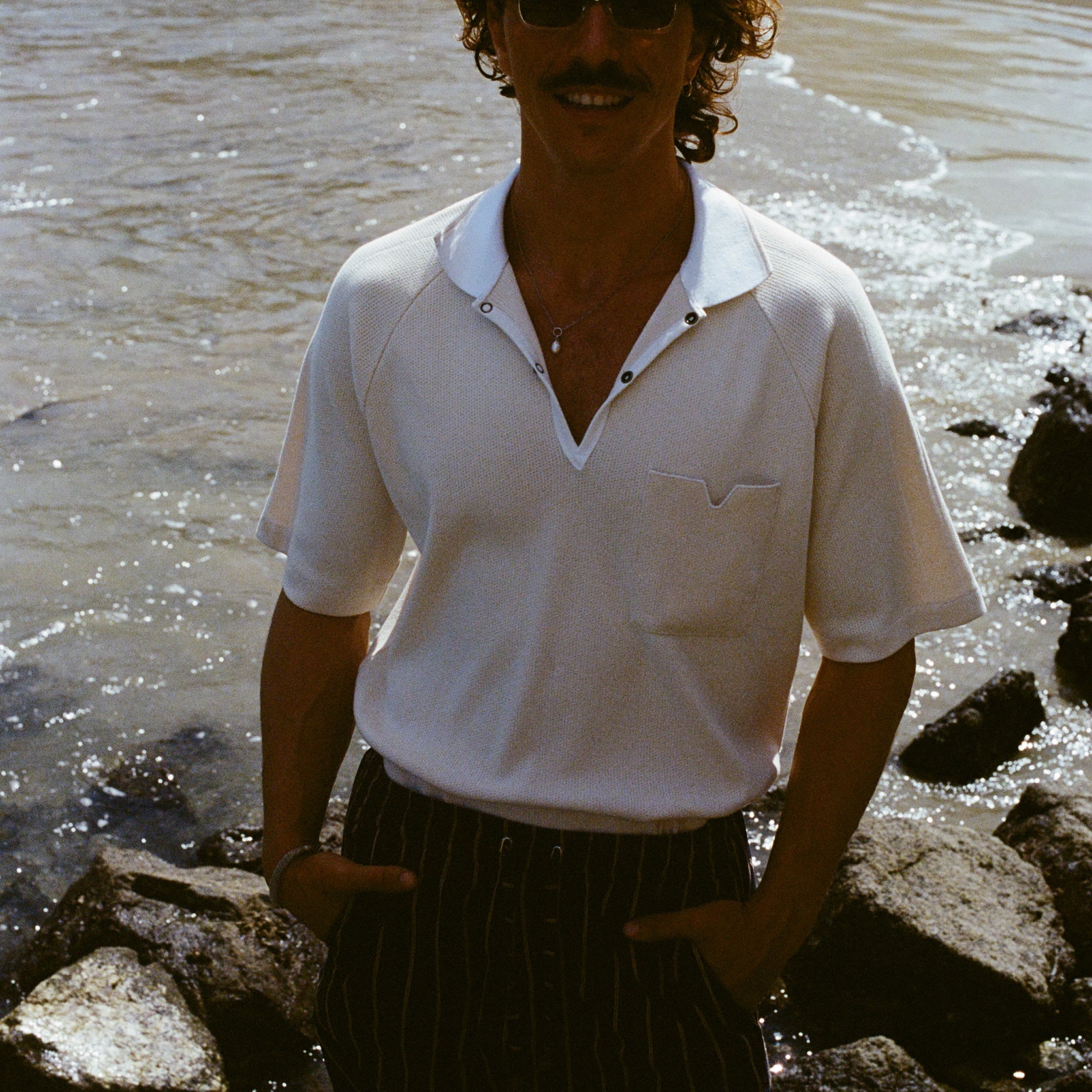 Man standing on rocky beach, wearing the beige mesh 'TomPolo' and striped shorts, and smiling to the camera with hands in pockets.