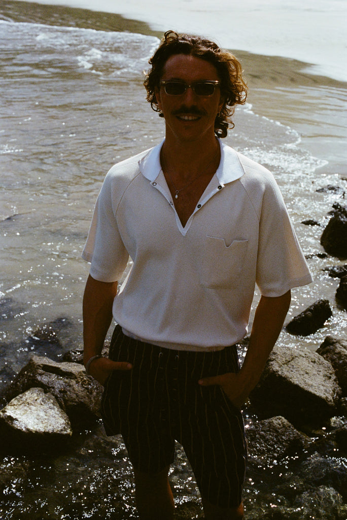 Man standing on rocky beach, wearing the beige mesh 'TomPolo' and striped shorts, and smiling to the camera with hands in pockets.