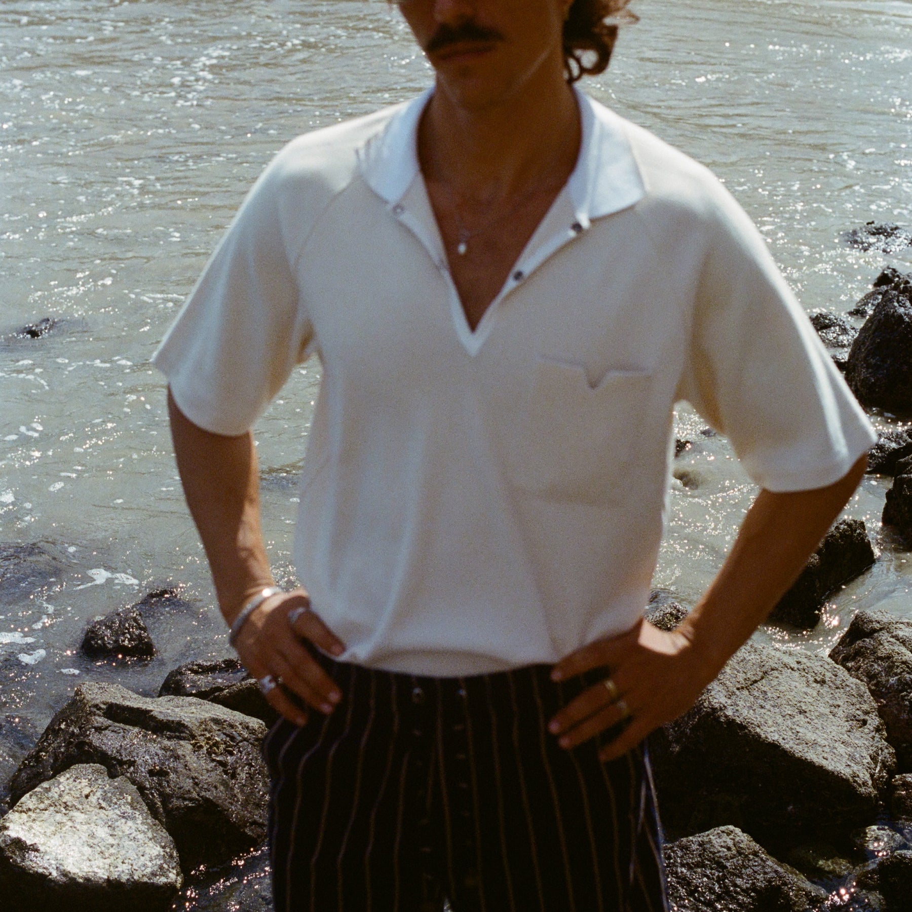Man standing on a rocky beach, wearing the striped Easy-Shorts and our beige mesh 'Tompolo'.