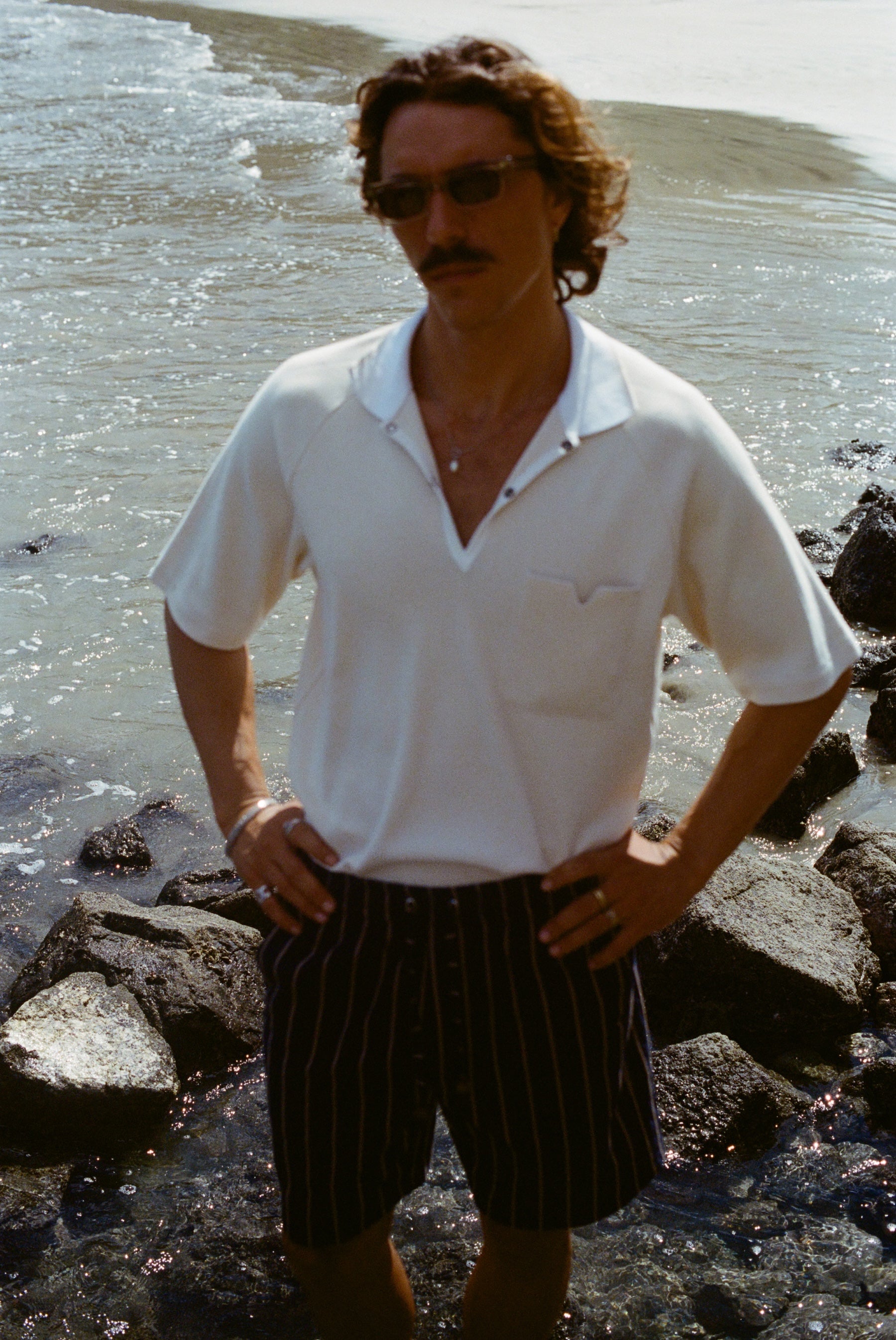 Man standing on a rocky beach, wearing the striped Easy-Shorts and our beige mesh 'Tompolo'.