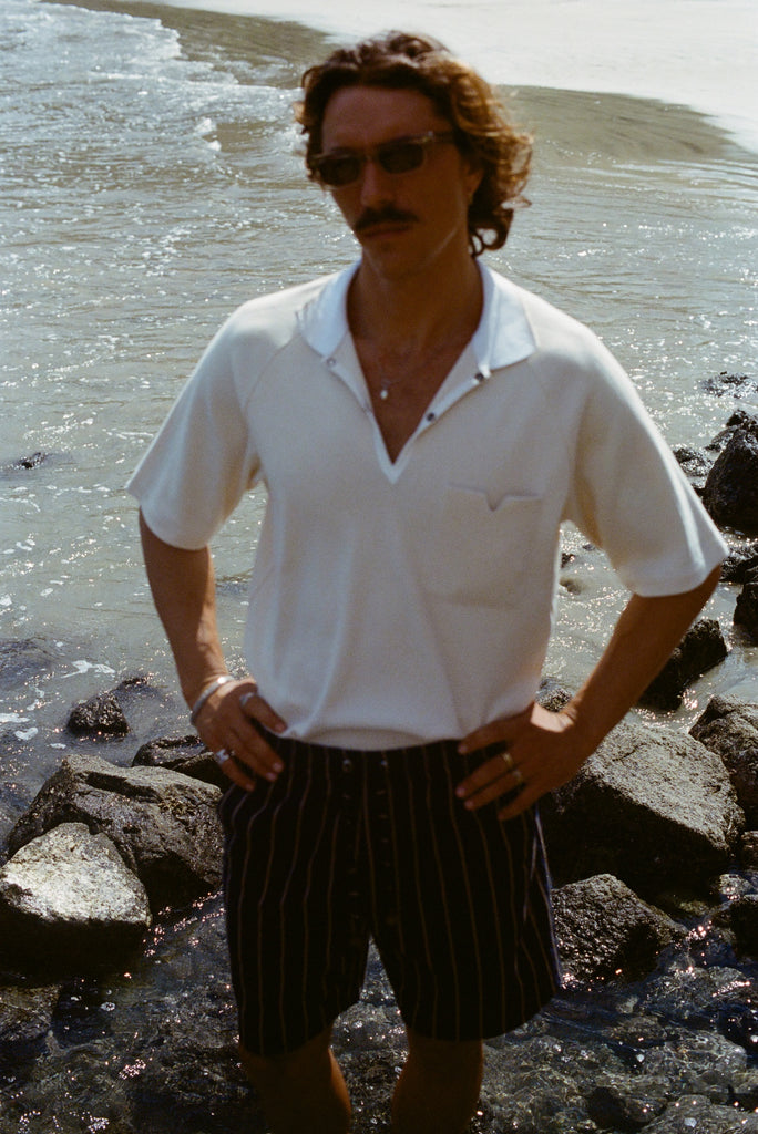 Man standing on a rocky beach, wearing the striped Easy-Shorts and our beige mesh 'Tompolo'.
