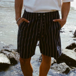 Close-up of a person standing on a shoreline wearing navy shorts with beige vertical stripes and a matching drawstring. The person is also wearing a white shirt with their hands in their pockets.