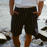 Close-up of a person standing on a shoreline wearing navy shorts with beige vertical stripes and a matching drawstring. The person is also wearing a white shirt with their hands in their pockets.