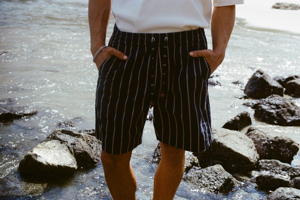 Close-up of a person standing on a shoreline wearing navy shorts with beige vertical stripes and a matching drawstring. The person is also wearing a white shirt with their hands in their pockets.