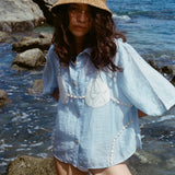 Image of woman in same light blue and dollar bikini shirt, standing ankle deep in the ocean, with a swim suit underneath and wearing a straw beach hat.