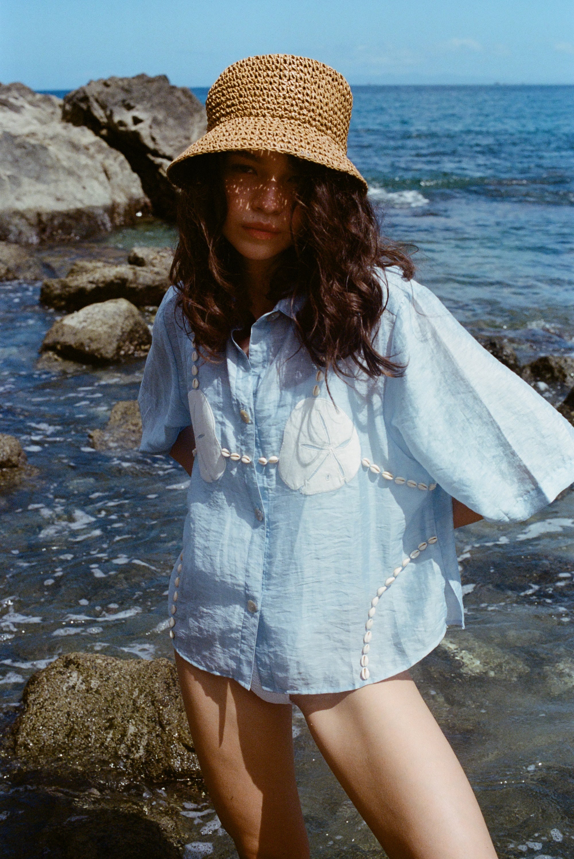 Image of woman in same light blue and dollar bikini shirt, standing ankle deep in the ocean, with a swim suit underneath and wearing a straw beach hat.