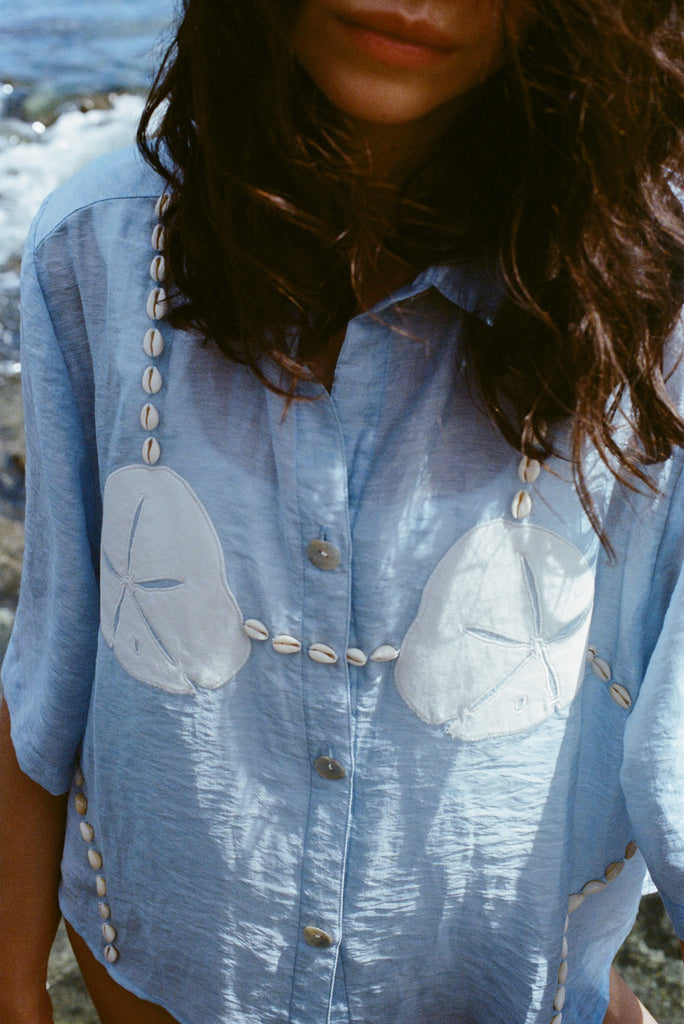 Close-up of a woman standing near the ocean wearing  light blue sand dollar bikini shirt.