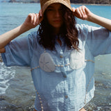 Image of woman in same light blue and dollar bikini shirt, standing ankle deep in the ocean, with a swim suit underneath and wearing a straw beach hat.