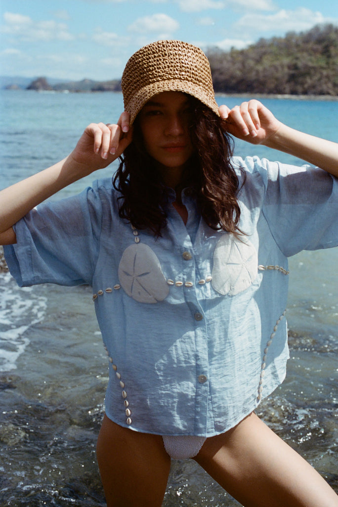 Image of woman in same light blue and dollar bikini shirt, standing ankle deep in the ocean, with a swim suit underneath and wearing a straw beach hat.