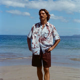 Male model posing on the beach with his hands on hips, wearing the linen brown shorts with a Tombolo Artist Button Down Shirt.