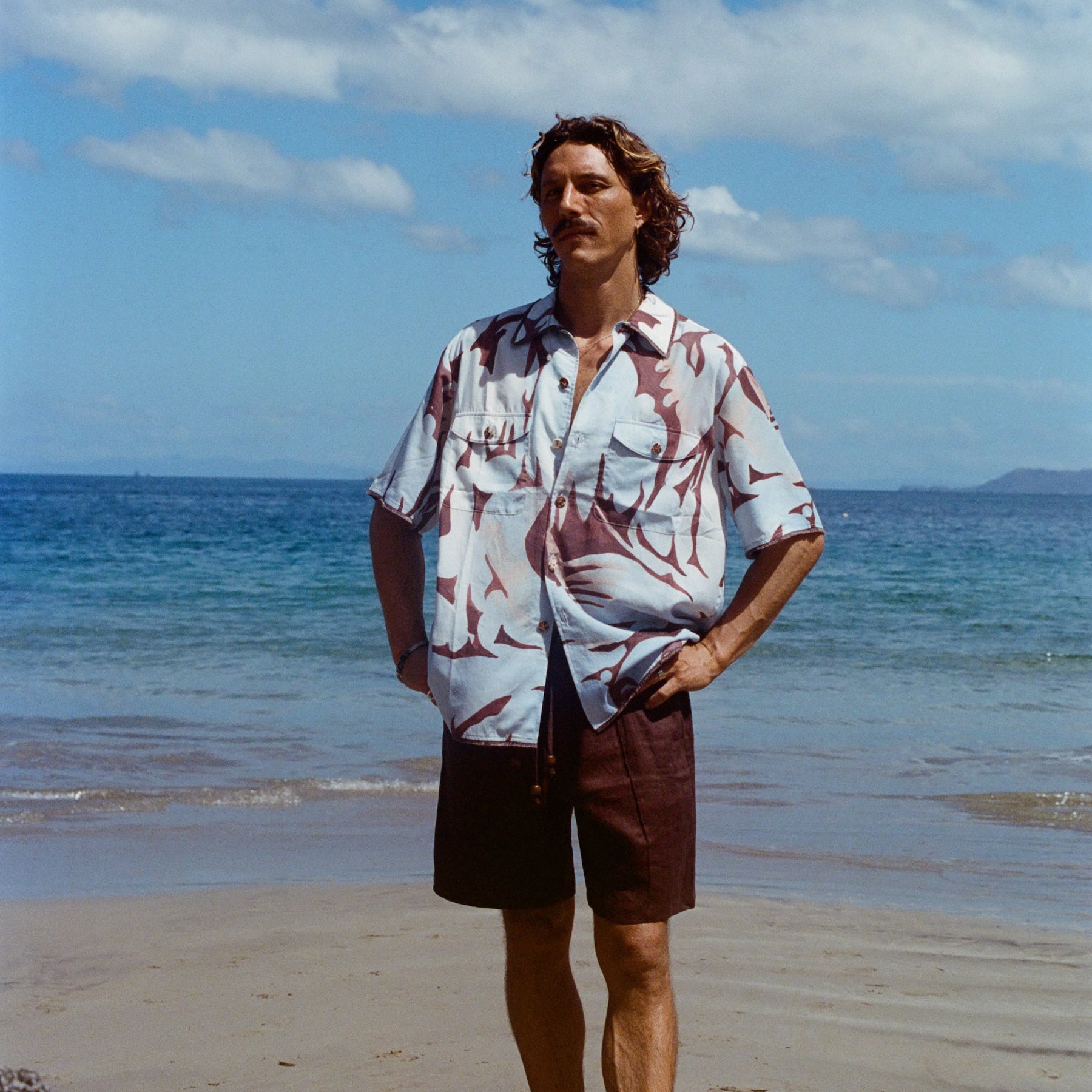 Male model posing on the beach with his hands on hips, wearing the linen brown shorts with a Tombolo Artist Button Down Shirt.