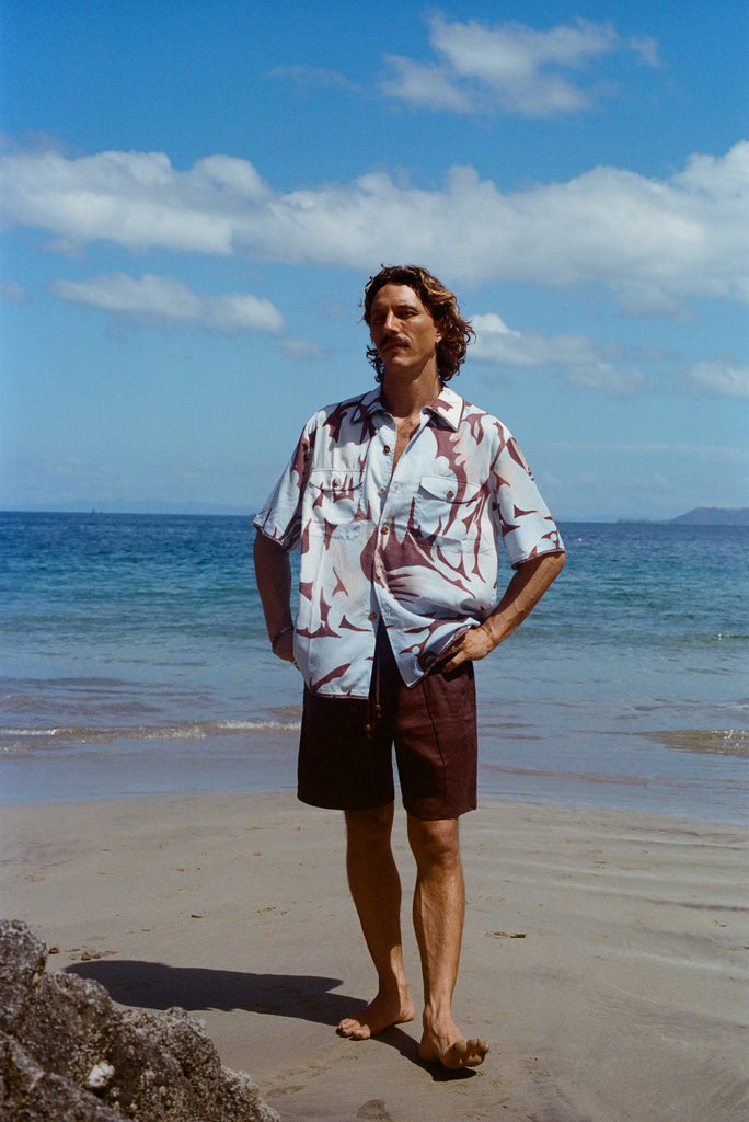 Male model posing on the beach with his hands on hips, wearing the linen brown shorts with a Tombolo Artist Button Down Shirt.