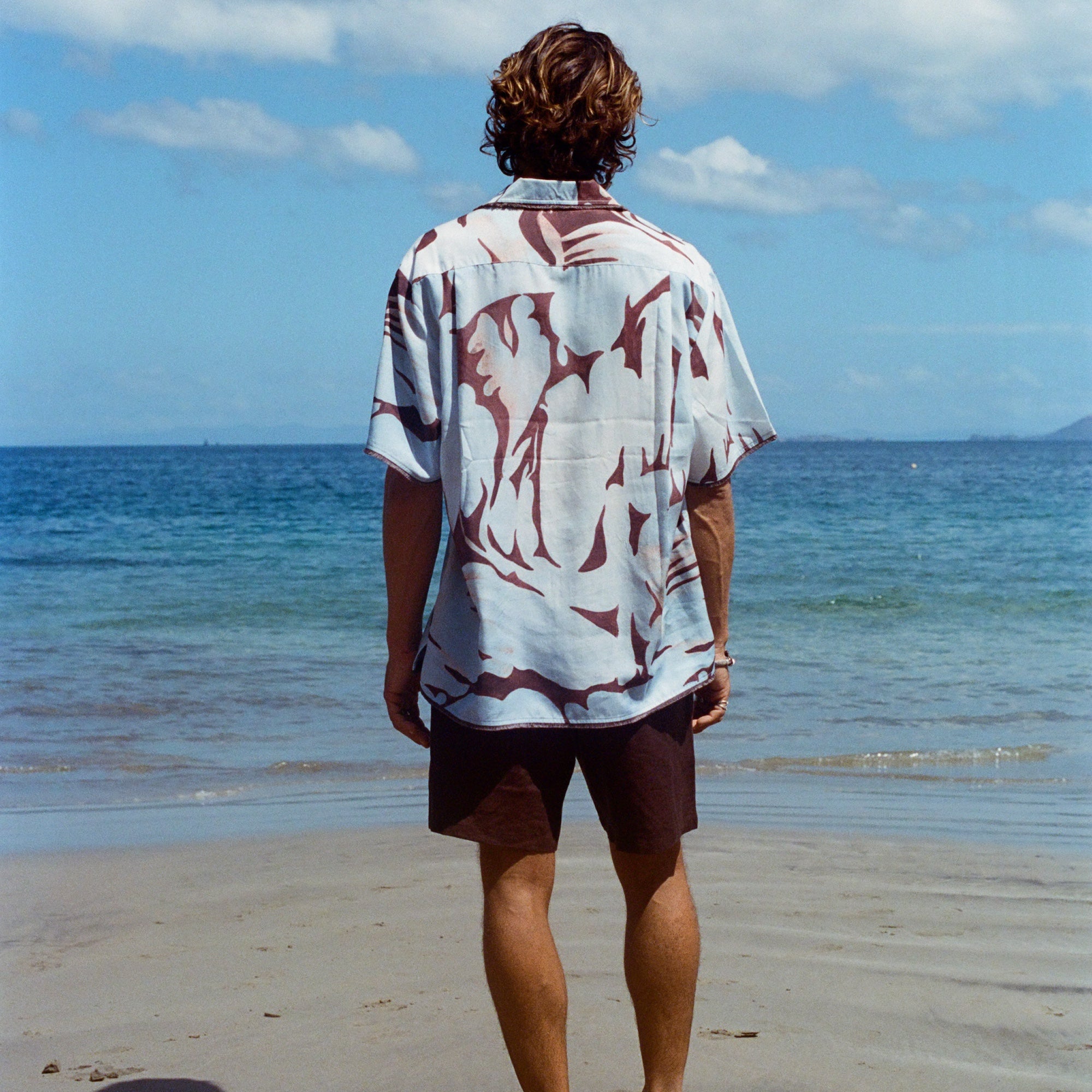 Image from behind of man on beach wearing the 'Mary Ball x Tombolo' Shirt with brown linen Easy Going Shorts.