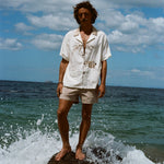 Male model pictured wearing the 'Monkey Business' Shirt with our Khaki Stowaway Shorts. He is standing on a rock at the beach with a wave splashing behind him.